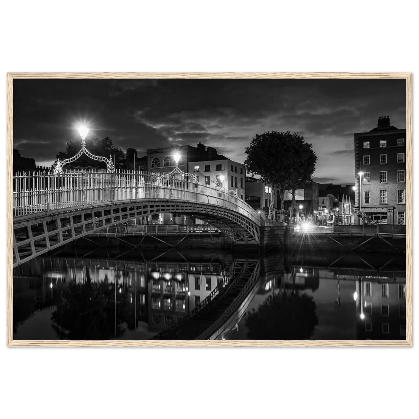  Black and white framed wall art print of the Ha'penny Bridge in Dublin at night. The bridge's historic architecture is beautifully highlighted against the dark sky, with reflections shimmering in the River Liffey below. Ideal for adding elegance to any room or office.